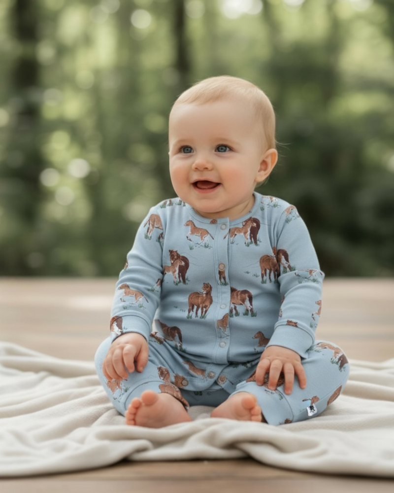 Smiling baby sitting on a cream blanket outdoors, dressed in a blue organic cotton romper featuring an all-over Exmoor pony print