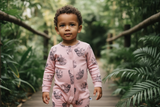 Toddler standing on wooden walkway surrounded by greenery, wearing pink sloth print organic cotton romper, Unisex